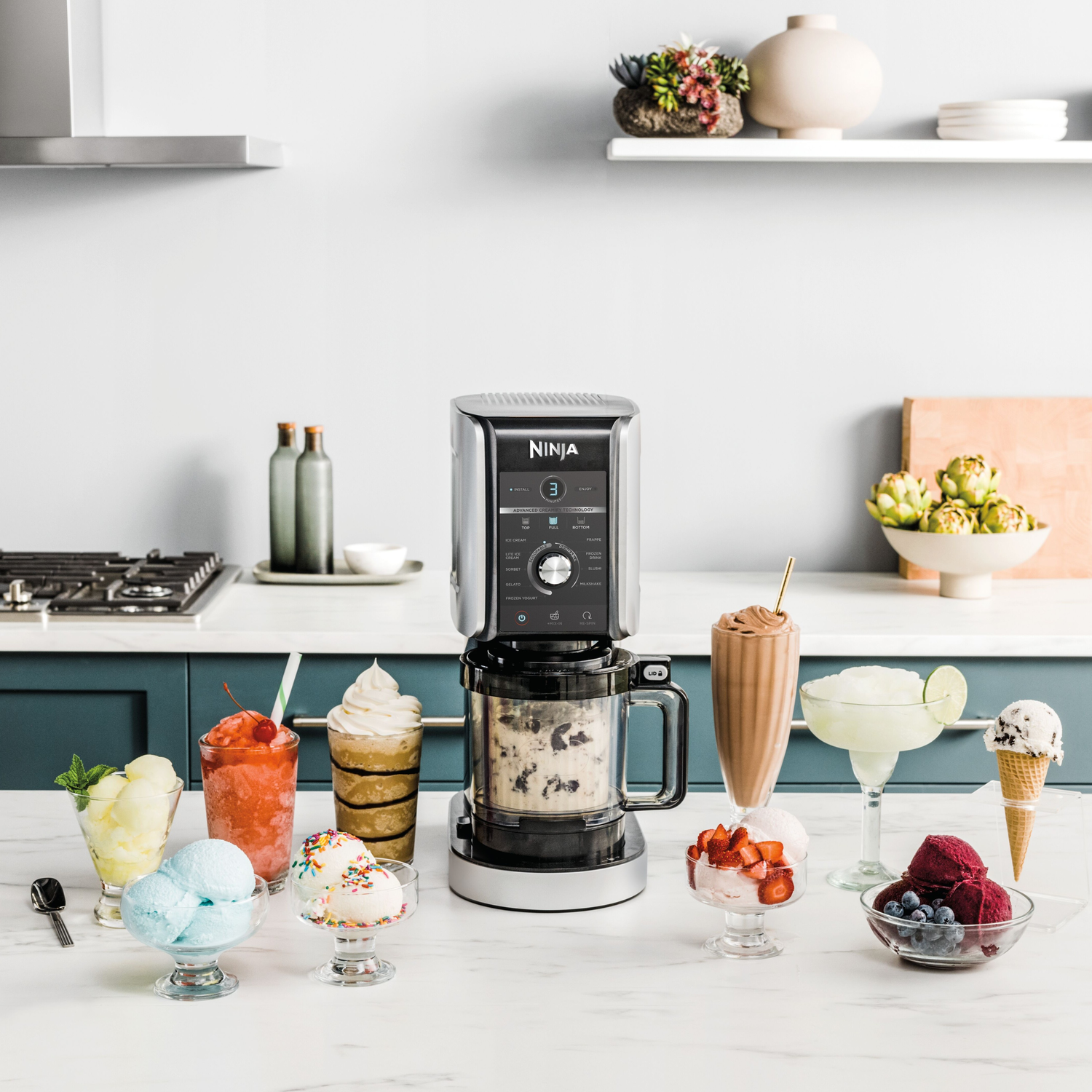 A Ninja blender with various ice cream desserts and drinks. Foreground: ice cream, smoothies. Background: kitchen, shelf with decor.