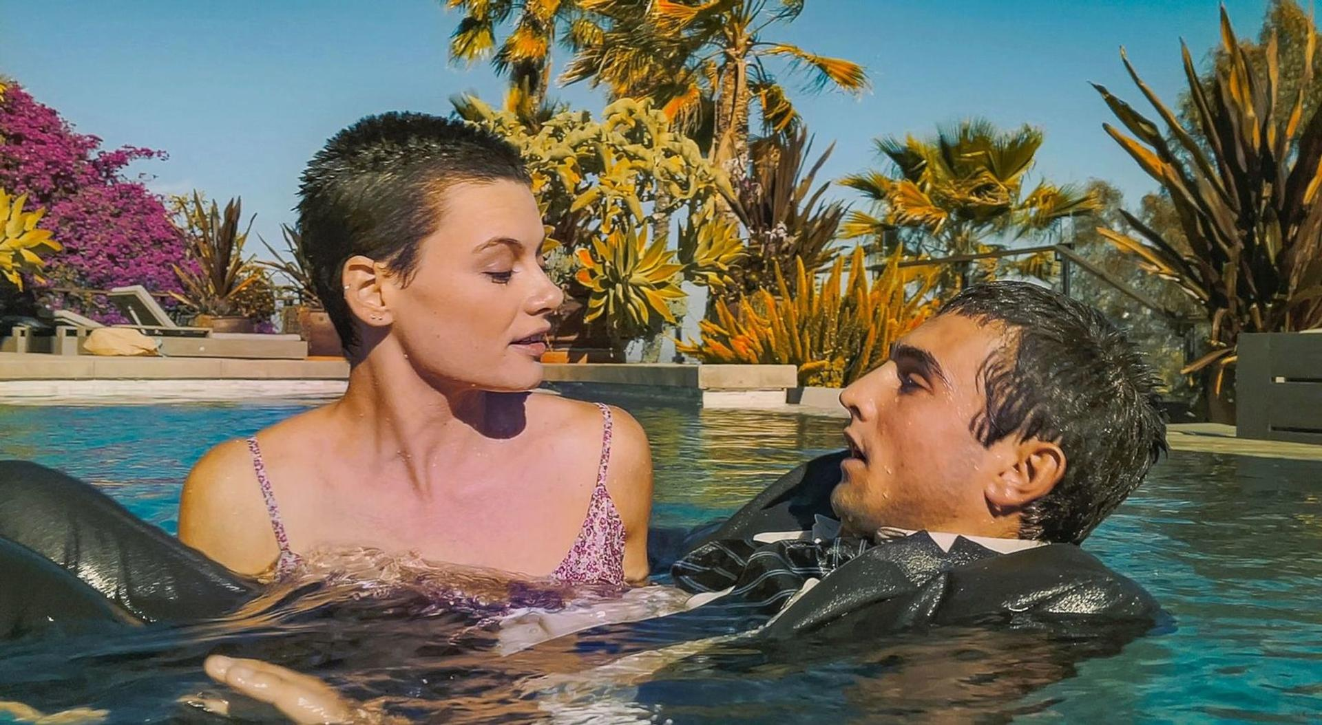 Eine Frau und ein Mann im Pool. Die Frau trägt ein rosa Oberteil. Der Mann, in einem Anzug, schwebt. Palmen und Blumen sind im Hintergrund.