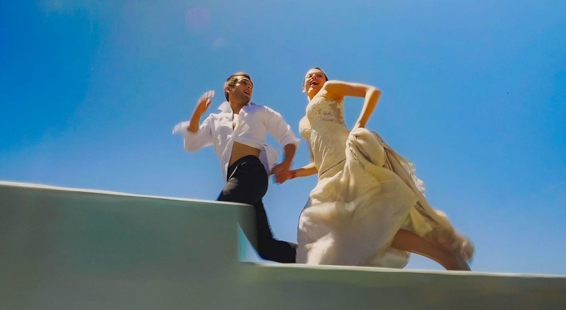Ein Paar in Hochzeitskleidung rennt unter blauem Himmel Stufen hinauf. Der Mann trägt ein weißes Hemd, die Frau ein Brautkleid.