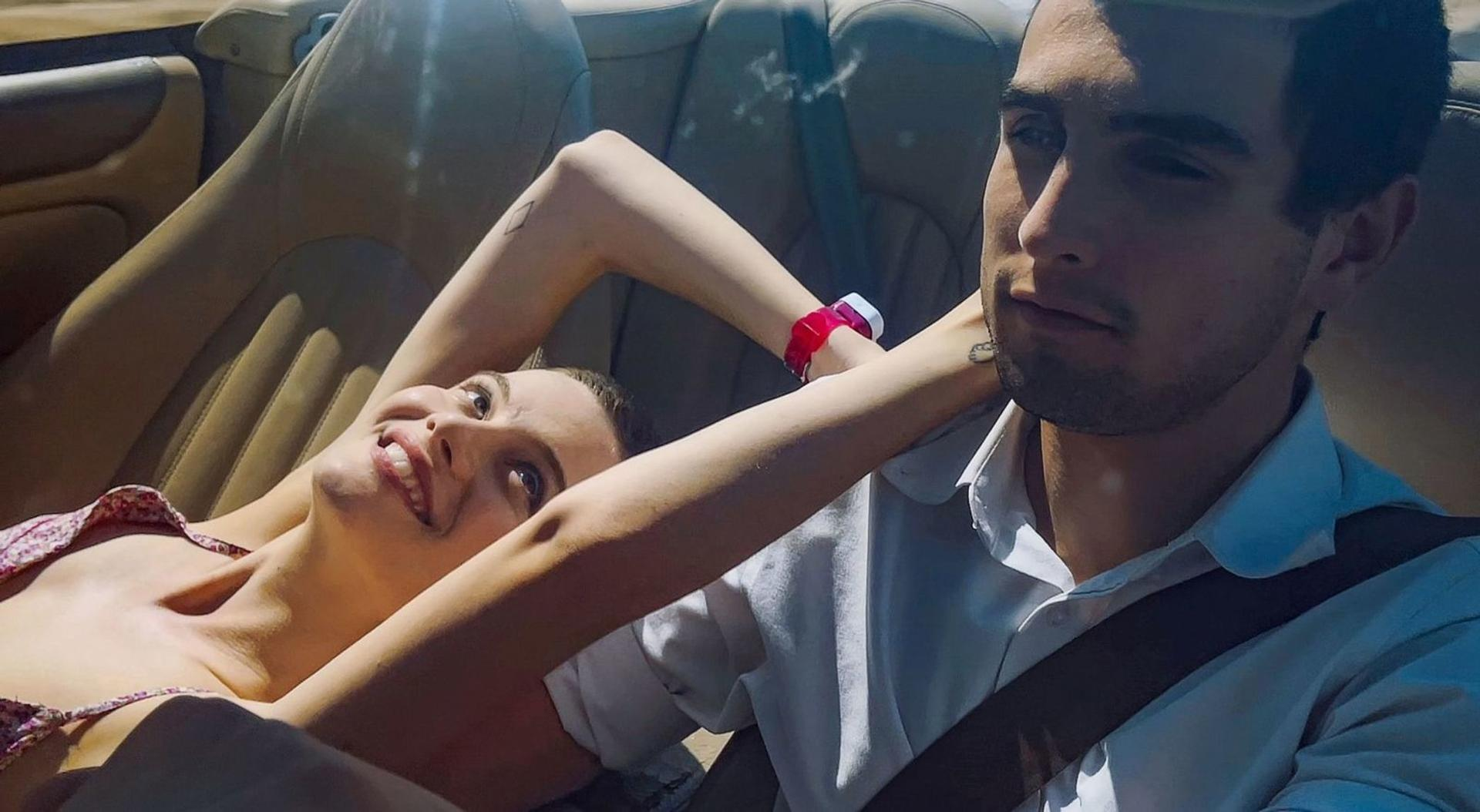 Eine Frau lächelt, liegt in einem Auto. Ein Mann schaut zu. Der Arm der Frau liegt auf der Schulter des Mannes.