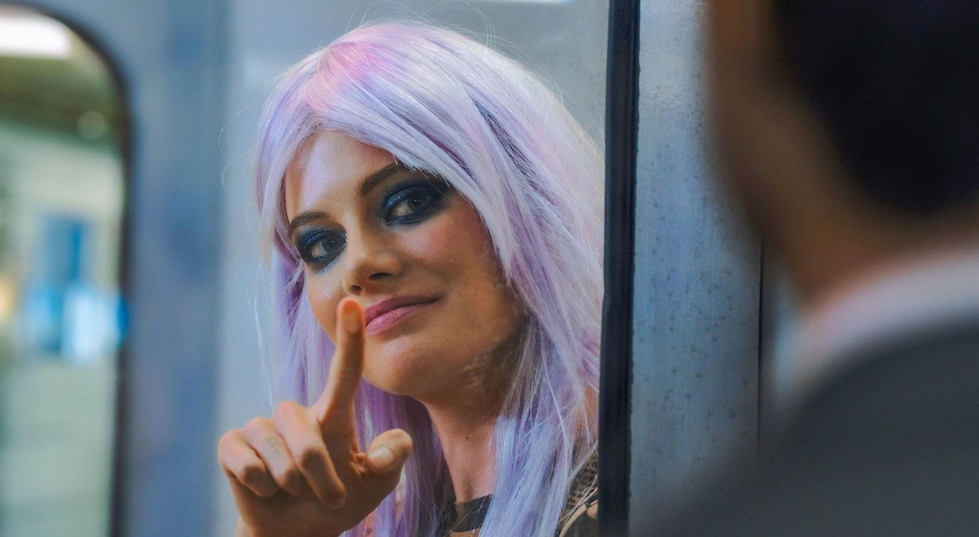 Frau mit lila Haaren, Finger an den Lippen, lächelnd. Eine andere Person ist im Hintergrund unscharf.