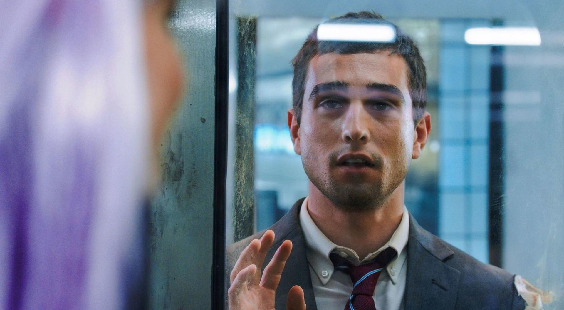Ein Mann im Anzug blickt durch Glas. Seine Hand ist auf dem Glas. Eine verschwommene Person ist auf der anderen Seite.