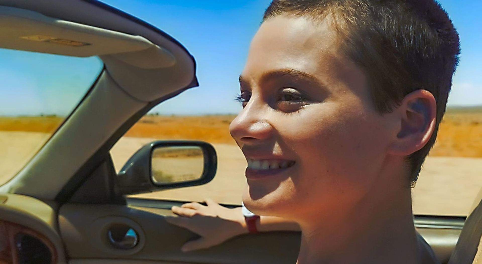 Eine Frau lächelt in einem Cabrio. Der Hintergrund zeigt eine Landschaft unter blauem Himmel. Innenraum und Seitenspiegel sind sichtbar.