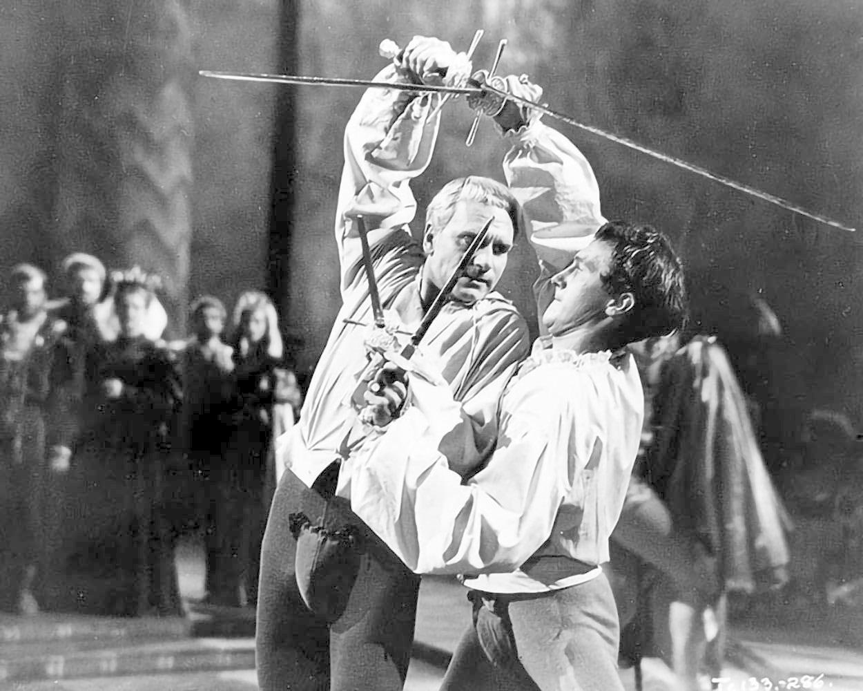 Zwei Männer in weißen Hemden duellieren sich mit Schwertern. Einer hält das Schwert hoch, der andere verteidigt sich. Zuschauer beobachten.