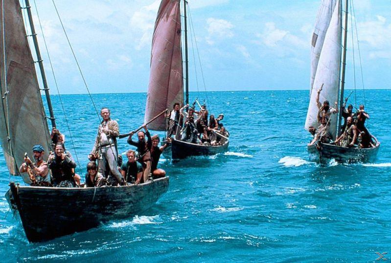 Drei Holzboote segeln auf dem Ozean. Jedes Boot trägt mehrere Personen mit gehissten Segeln. Der Himmel ist blau.