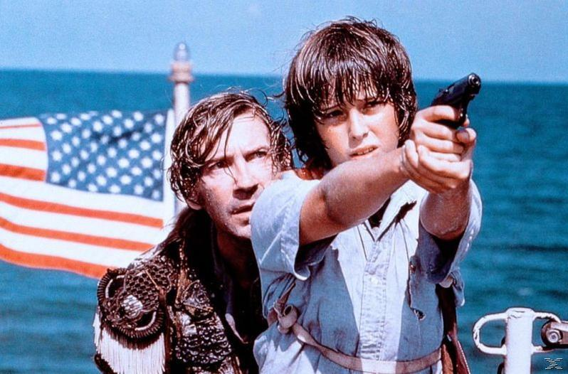 Zwei Personen auf einem Boot. Einer hält eine Waffe und zielt nach vorne. Eine amerikanische Flagge und das Meer sind im Hintergrund.