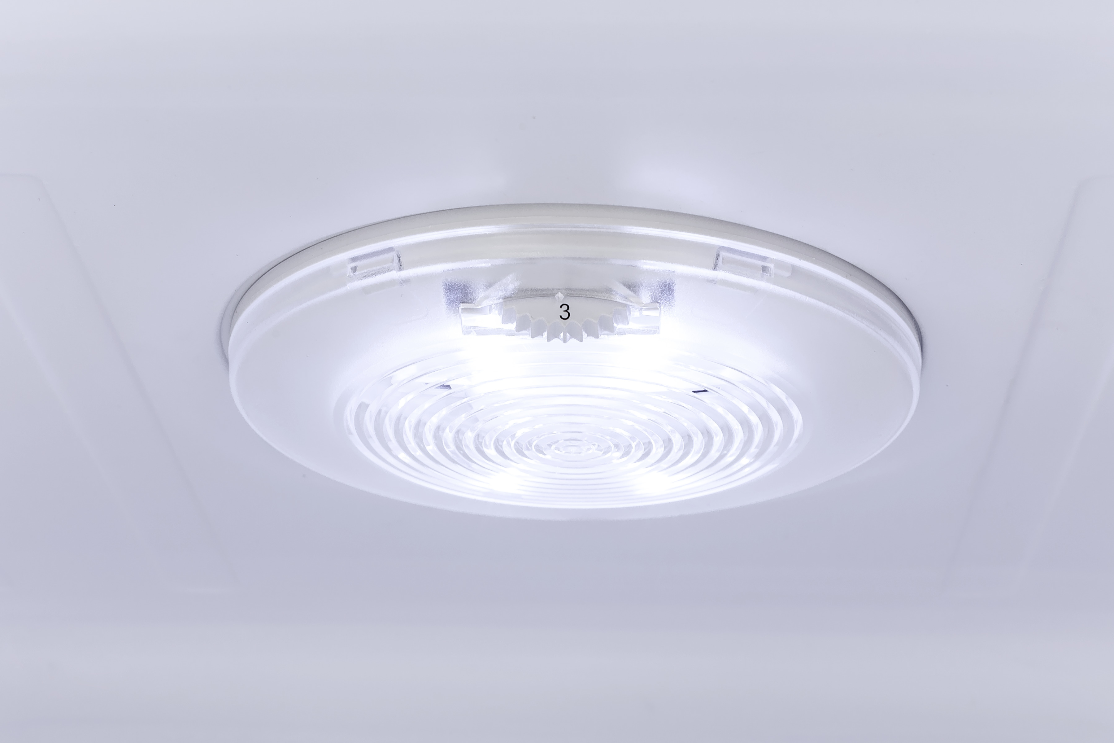 Nahaufnahme einer weißen, kreisförmigen Leuchte an einer weißen Decke. Die Leuchte hat eine Zahl 3 und ein geriffeltes Innendesign.