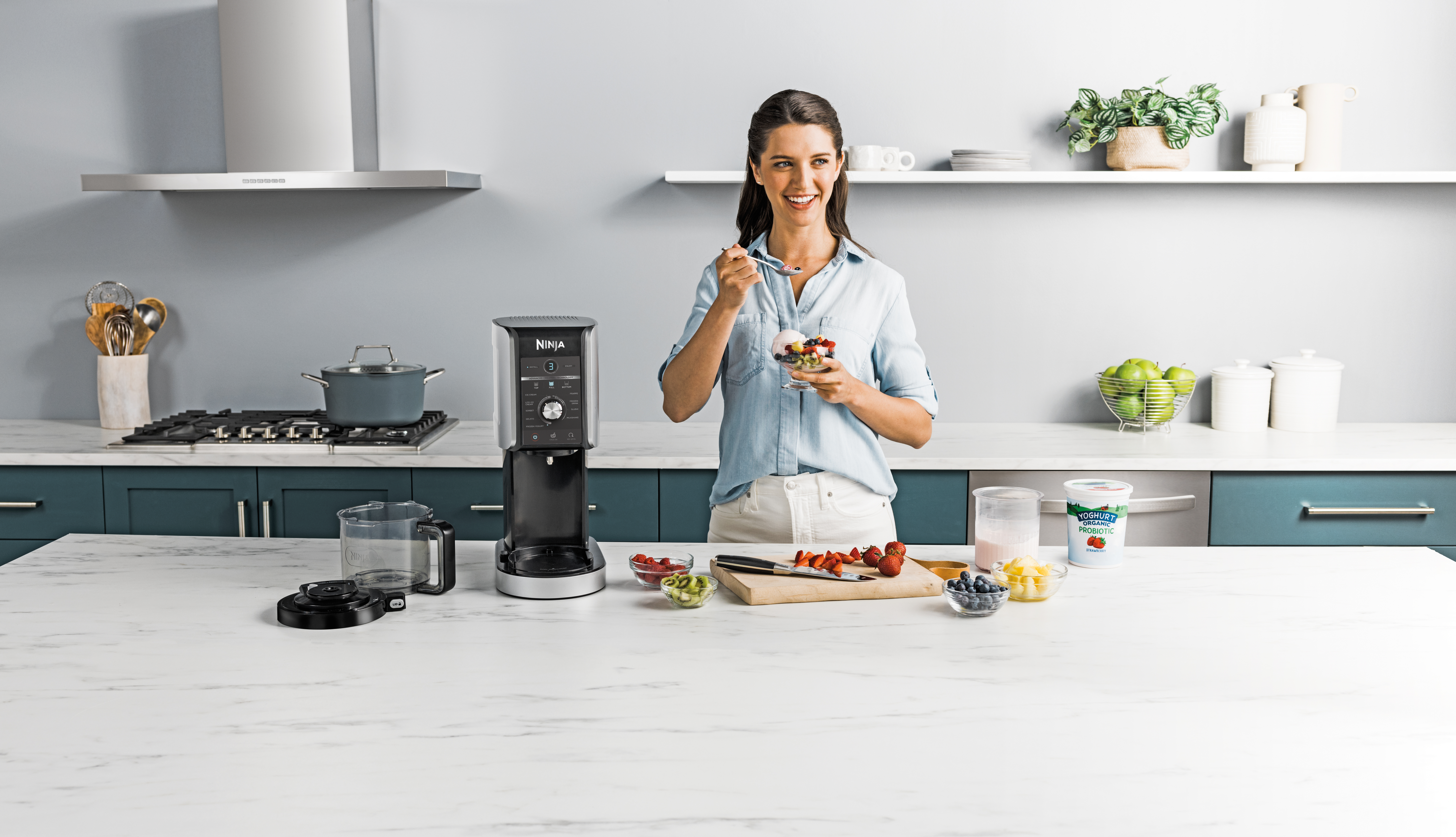 Femme dans une cuisine mangeant des fruits. Une cafetière Ninja et des ingrédients sur le comptoir.