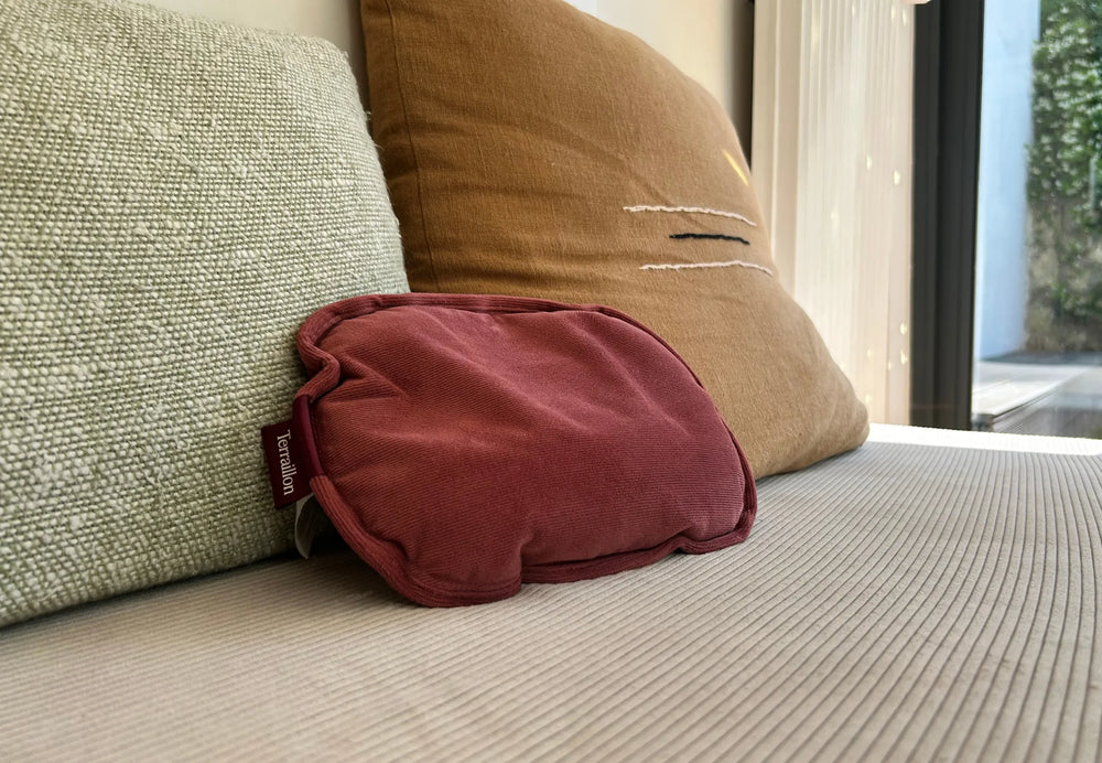 Un oreiller bordeaux repose sur un coussin côtelé. Deux autres oreillers, un vert et un beige, sont derrière. Une fenêtre est visible.