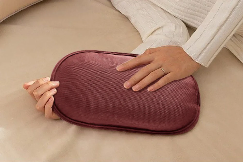 Une personne tient un oreiller bordeaux. Les mains de la personne sont visibles, avec une bague au doigt. Le fond est un lit.