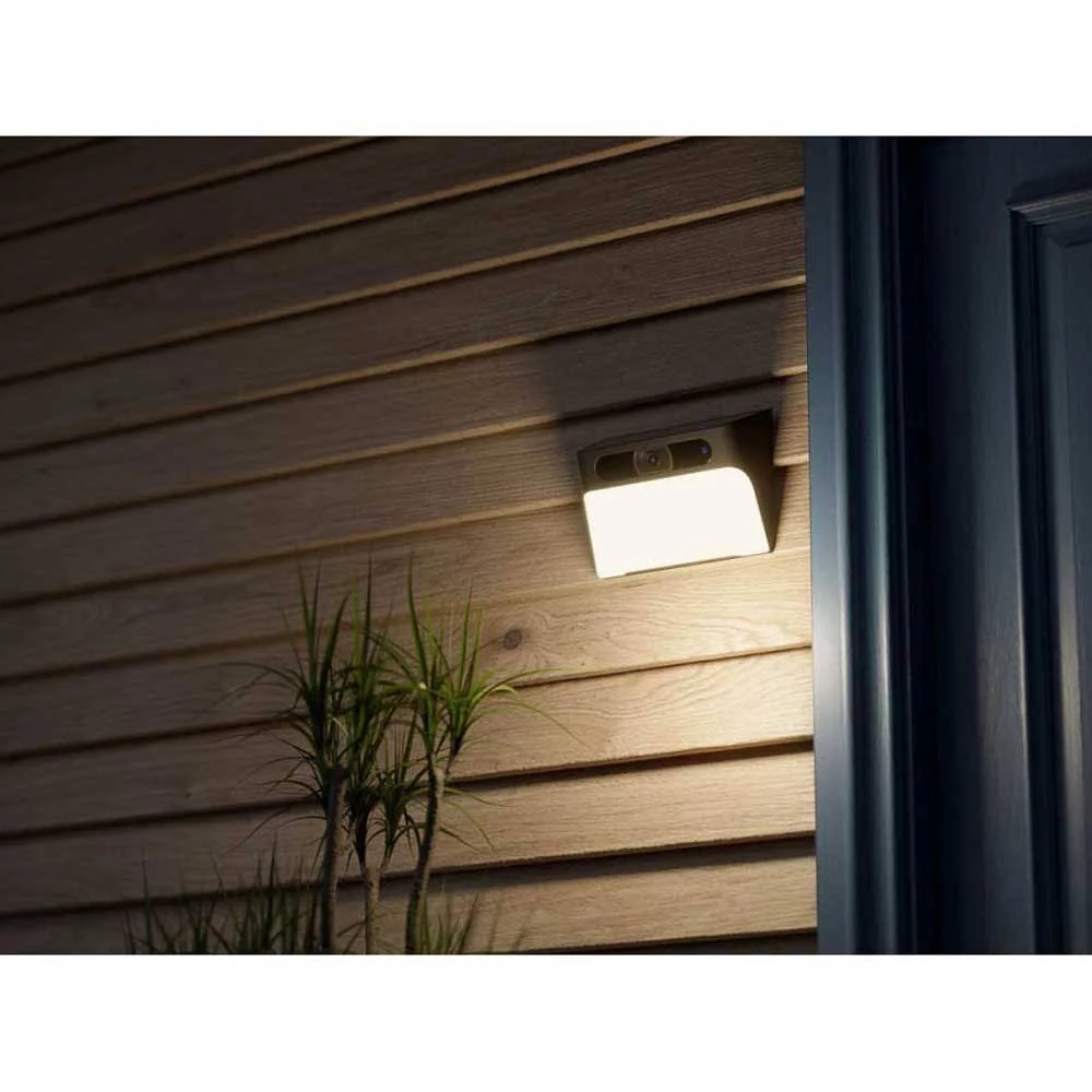 Una lámpara exterior está montada en un revestimiento de madera. Hay una pequeña planta delante. La luz está encendida, iluminando el área.