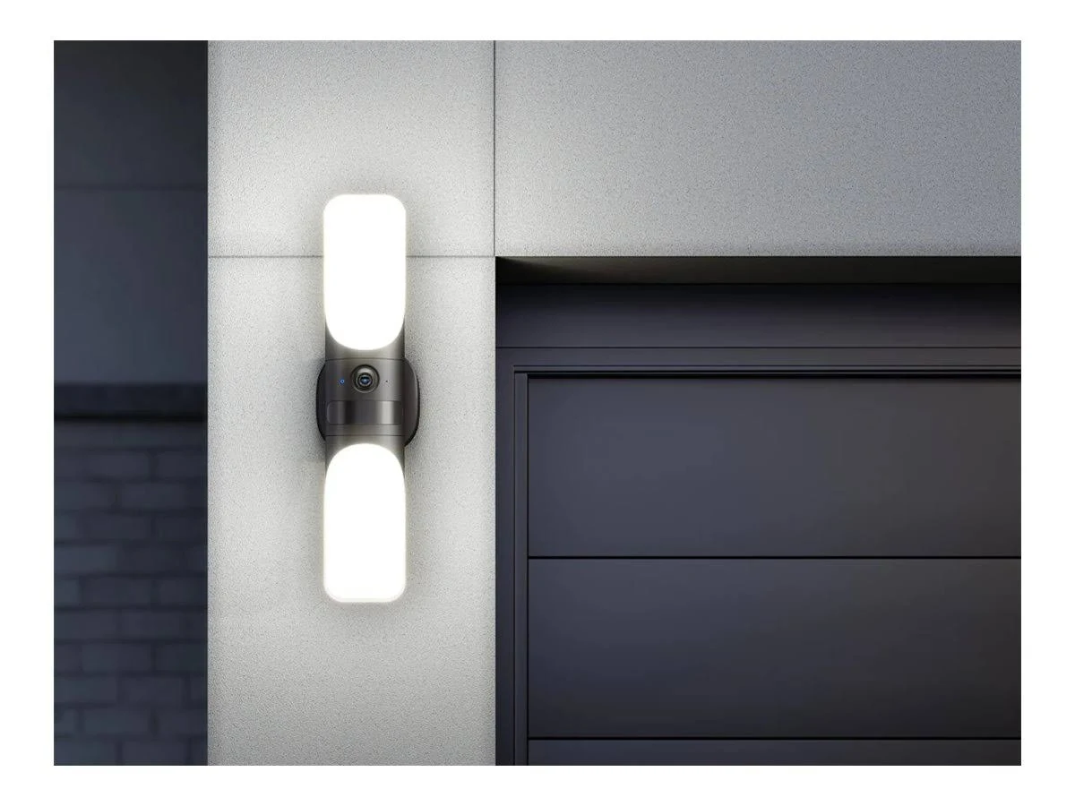 Una luz de seguridad montada en la pared con dos luces brillantes y una cámara. El fondo tiene una puerta oscura y una pared texturizada.