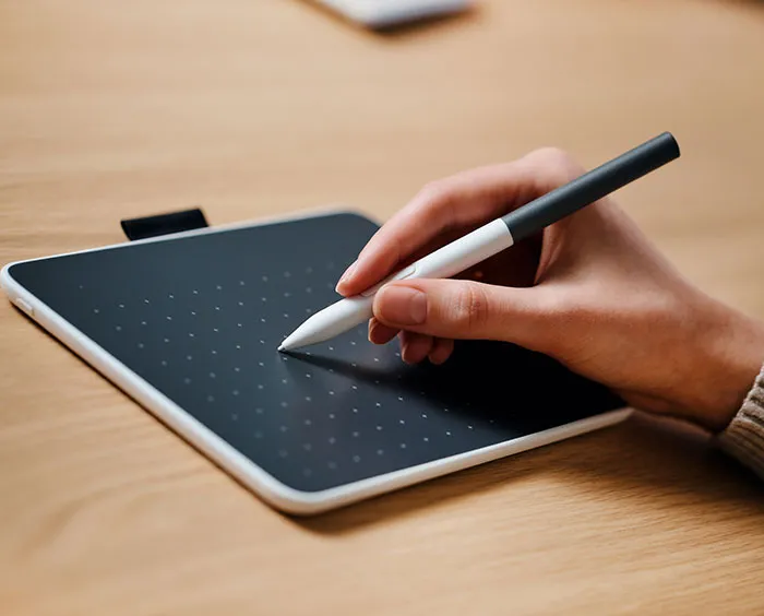 Una mano tiene una penna bianca sopra un tablet nero con puntini. Lo sfondo mostra un tavolo di legno.