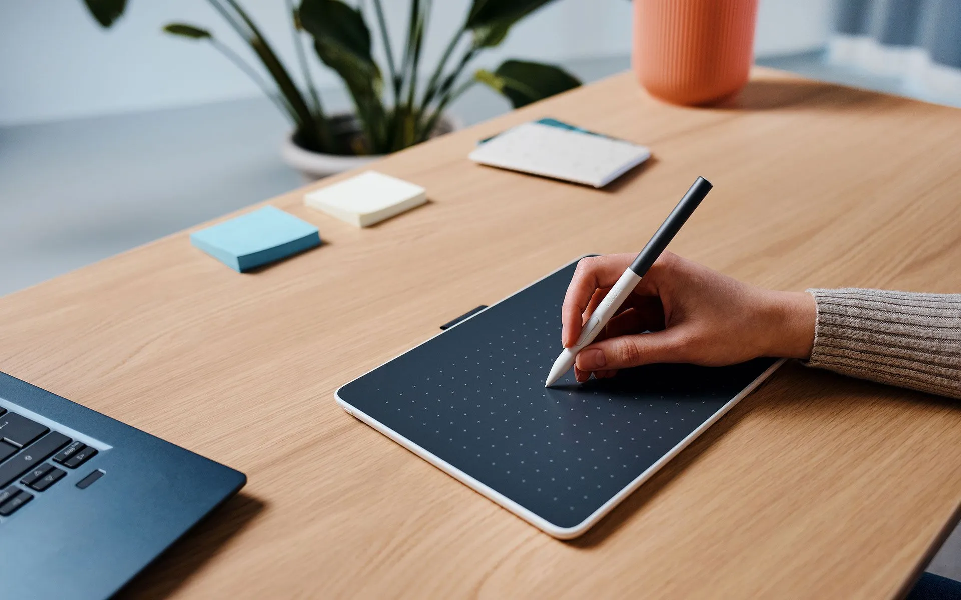 Une main tenant un stylo au-dessus d'une tablette graphique. Un ordinateur portable, des post-it et une plante sont sur la table en bois.