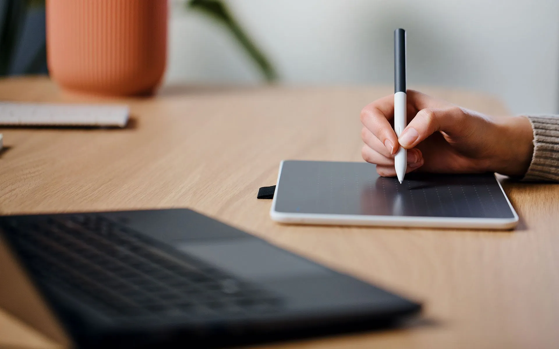 Une main tenant un stylo au-dessus d'une tablette graphique. Un ordinateur portable est au premier plan. Un pot est en arrière-plan.