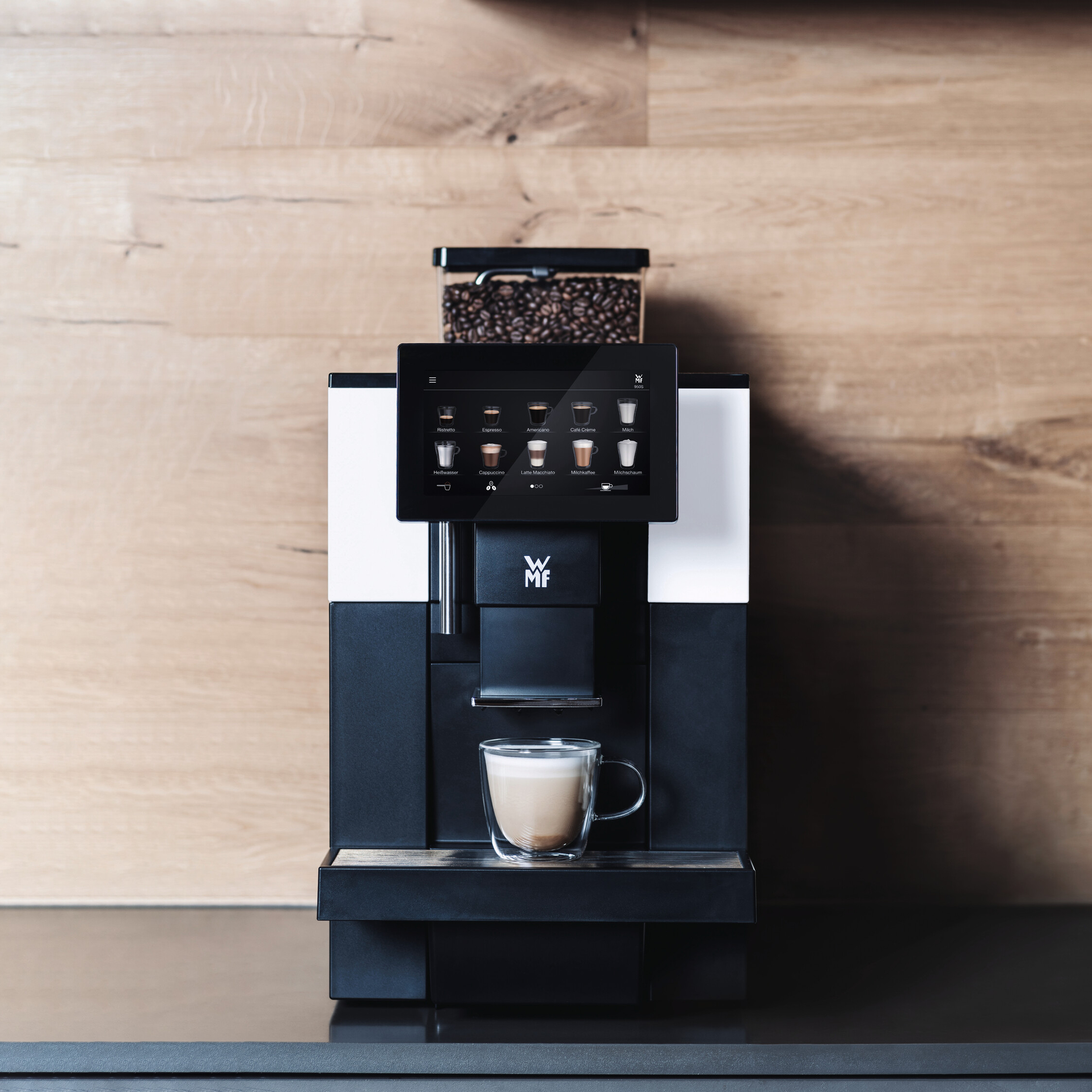 Eine Kaffeemaschine mit Touchscreen, der Kaffeeoptionen anzeigt. Ein Glas Latte steht unten. Holz Hintergrund.