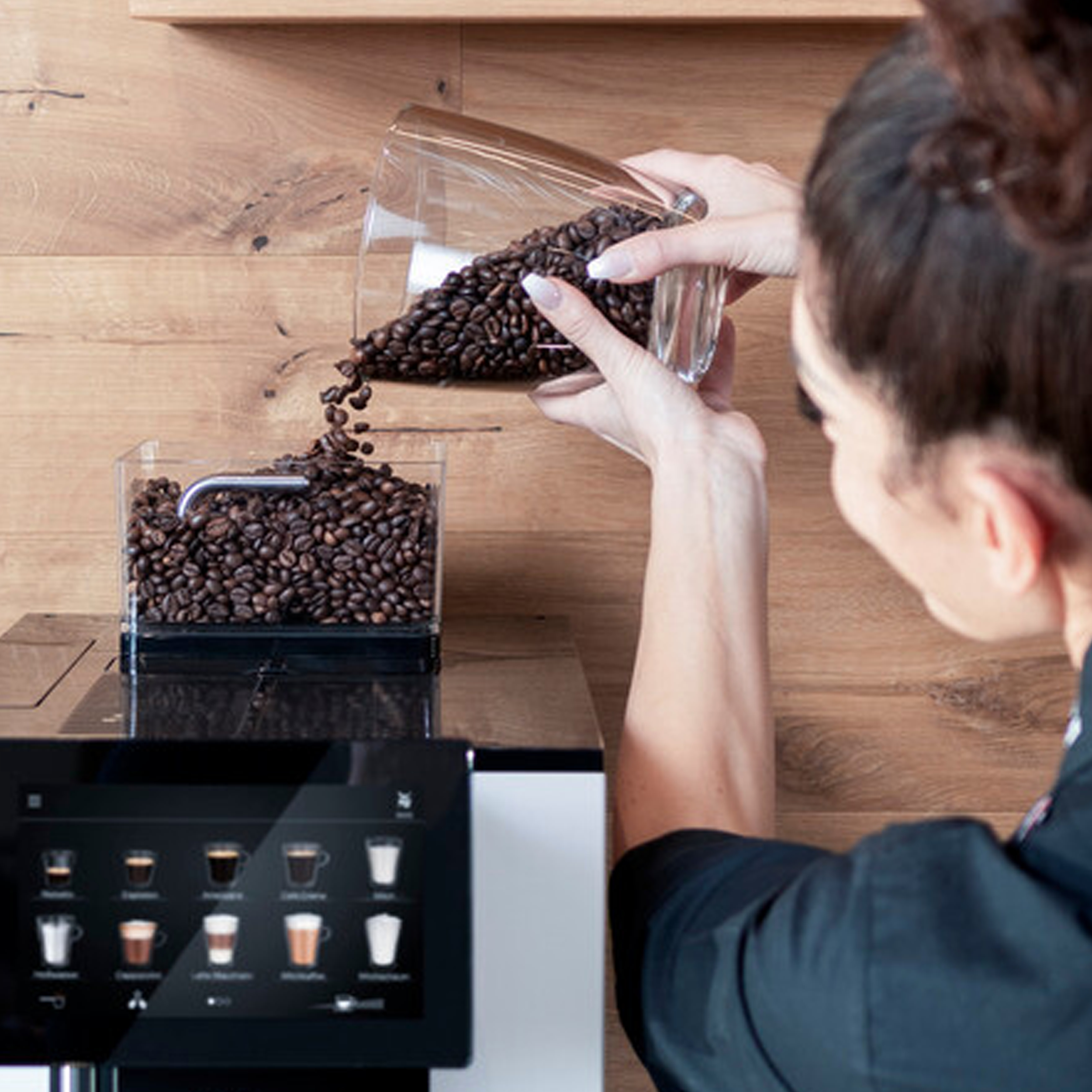 Eine Person gießt Kaffeebohnen aus einem Behälter in eine Kaffeemaschine. Die Maschine hat einen Touchscreen.