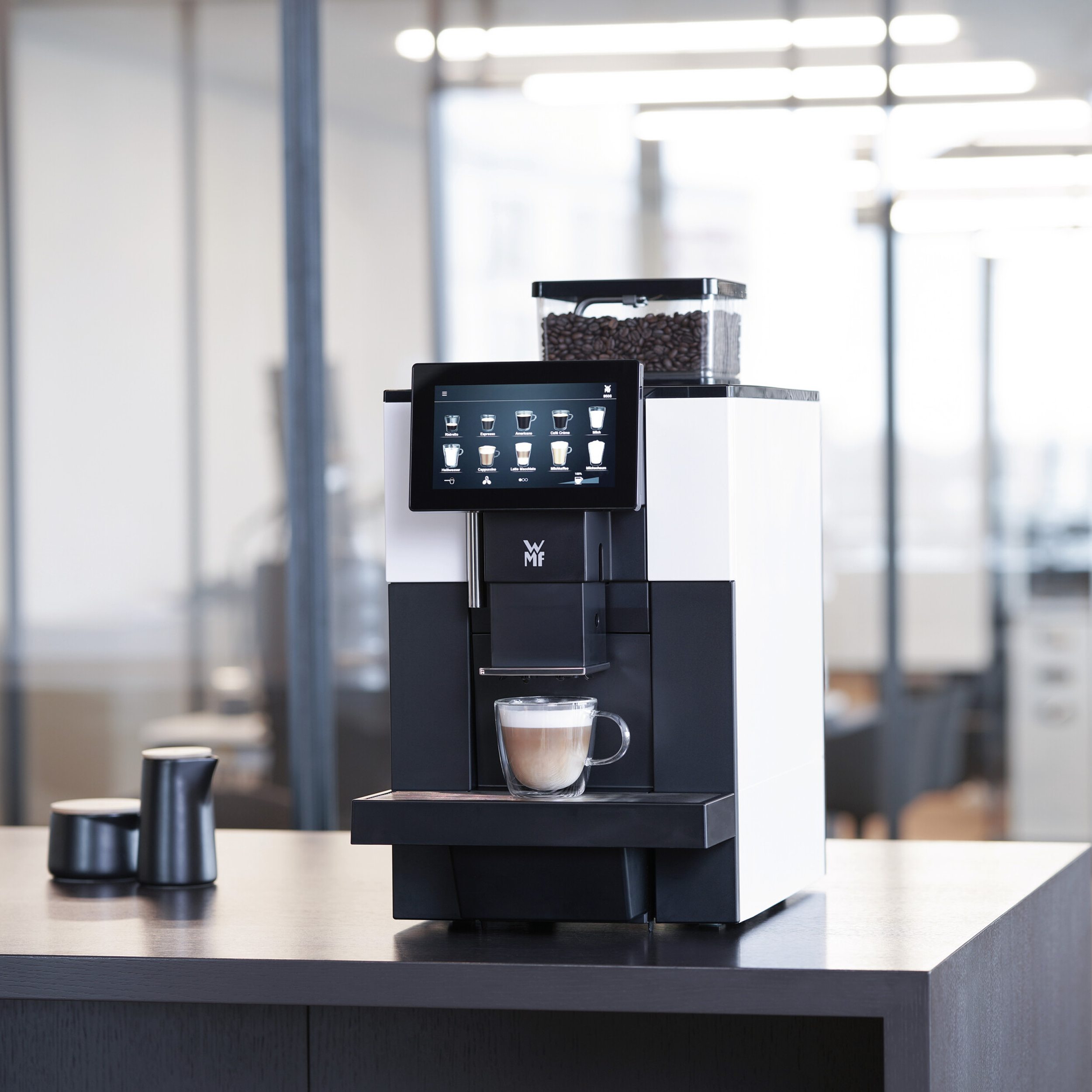 Eine Kaffeemaschine mit Touchscreen, Kaffeebohnenbehälter und einem Glas mit Kaffee. Zwei schwarze Behälter links.