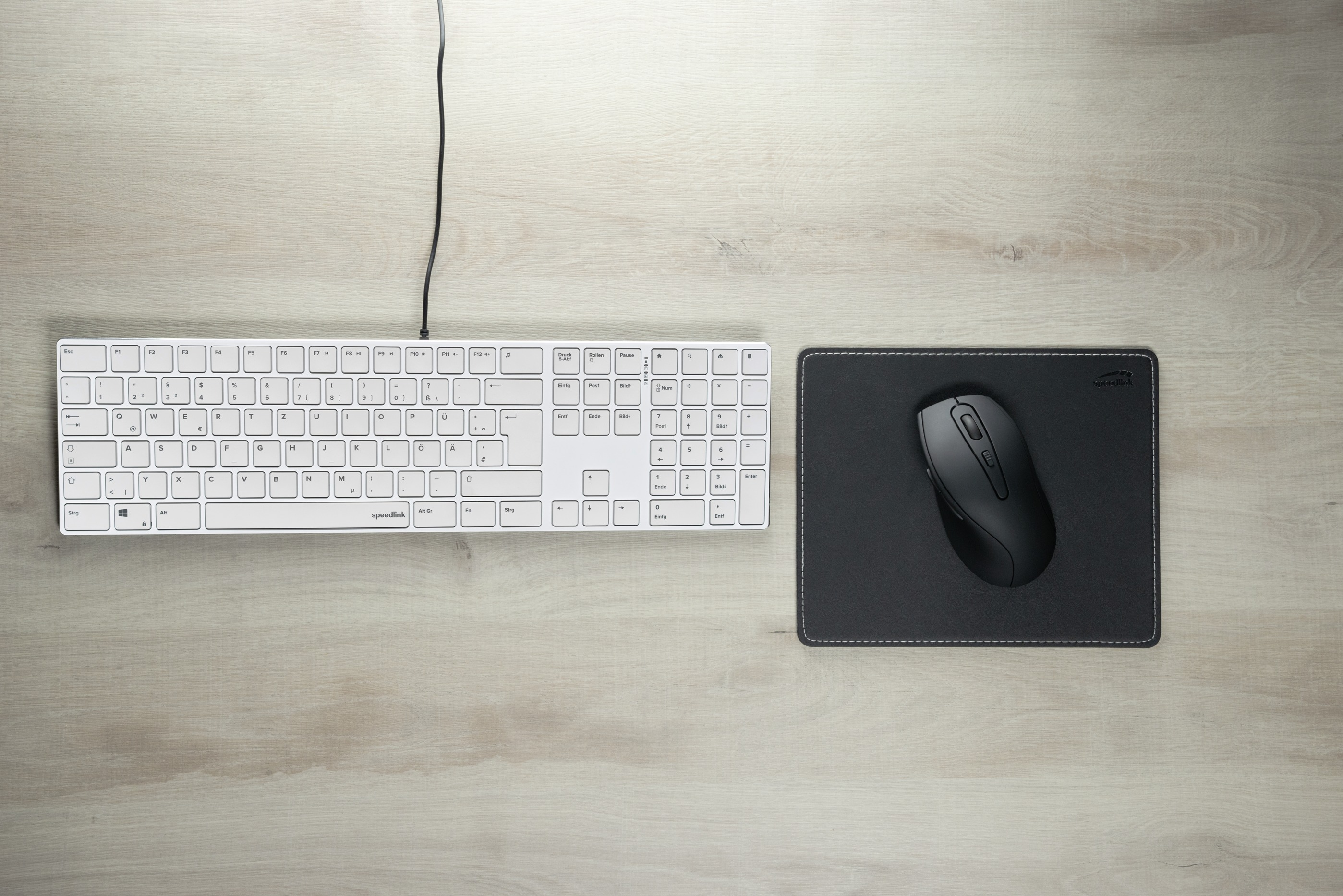 Eine weiße Tastatur und eine schwarze Maus auf einem Holztisch. Ein schwarzes Mauspad ist auch vorhanden.