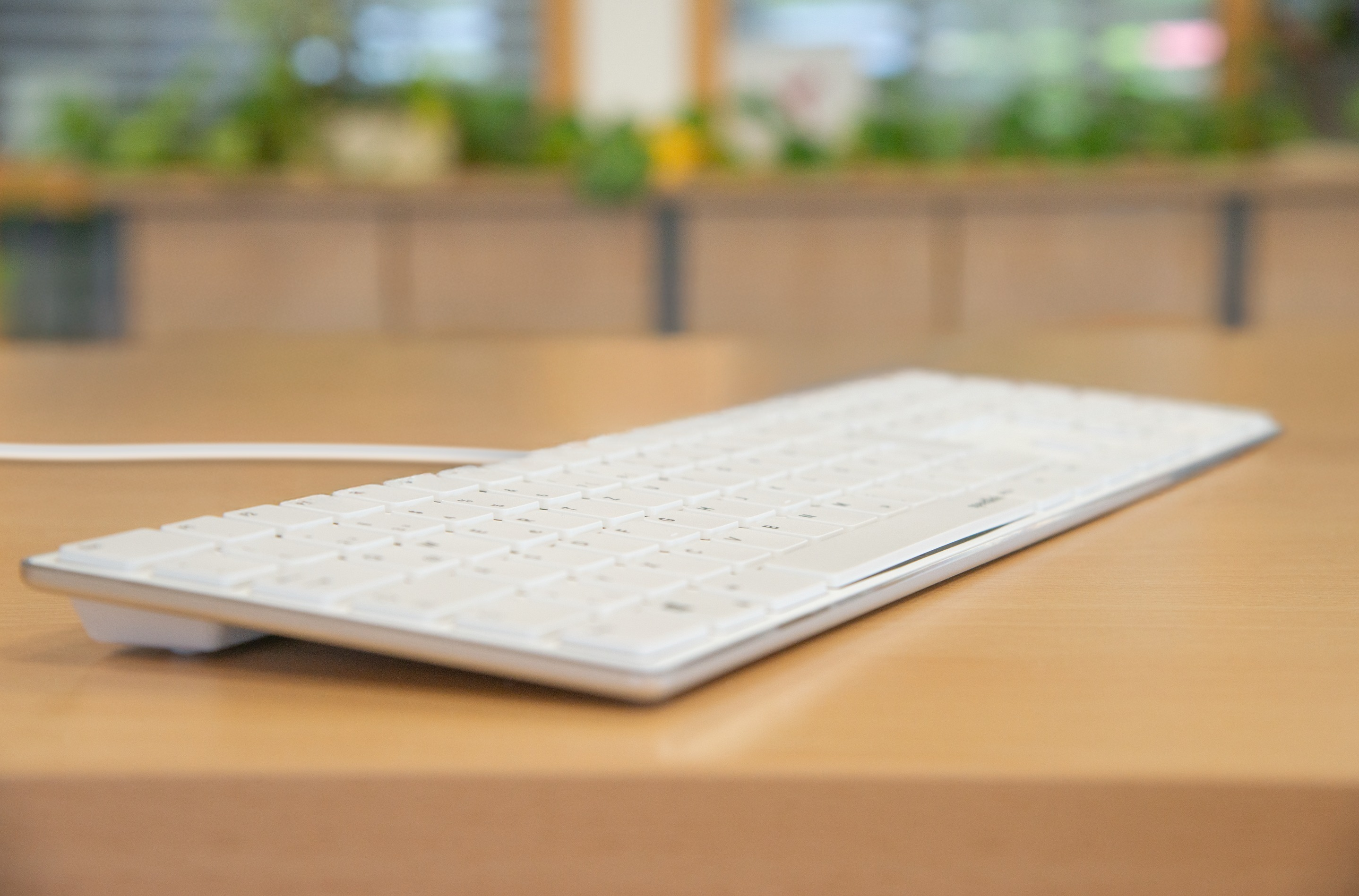 Eine weiße Tastatur auf einem hellbraunen Holztisch. Der Hintergrund ist unscharf und zeigt Grün und eine hellbraune Struktur.