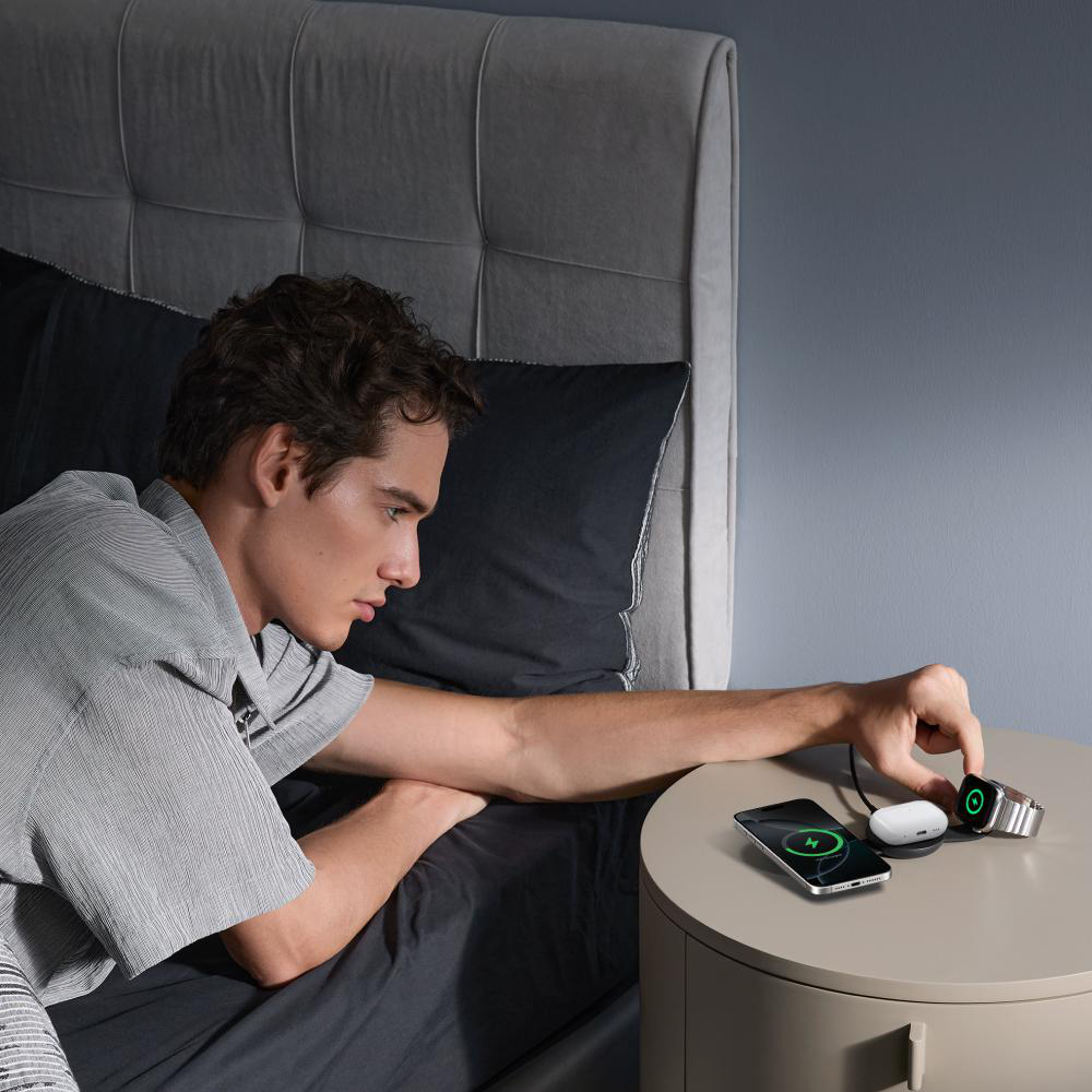 Mann im Bett greift nach Smartwatch auf Nachttisch. Telefon und Ohrhörer werden ebenfalls geladen. Schlafzimmer mit dunkler Bettwäsche.
