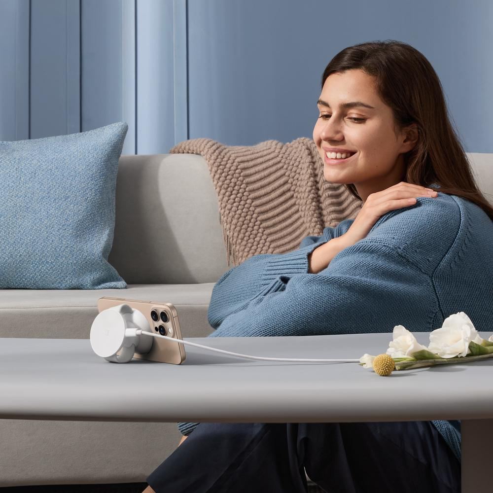 Eine lächelnde Frau blickt auf ein Telefon, das auf einem Tisch geladen wird. Hinter ihr sind ein Sofa und ein Kissen. Blumen sind auf dem Tisch.