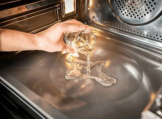 Osoba wlewa wodę ze szklanki do piekarnika. Woda rozlewa się po metalowej powierzchni piekarnika.