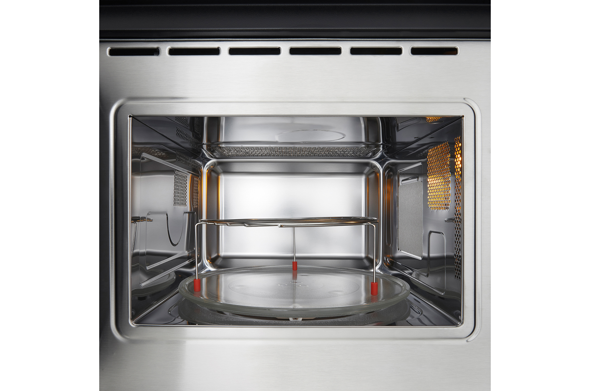 Interior de microondas abierto con plato giratorio de vidrio y rejilla. Interior de acero inoxidable brillante con ventilación arriba. Iluminado a la derecha.