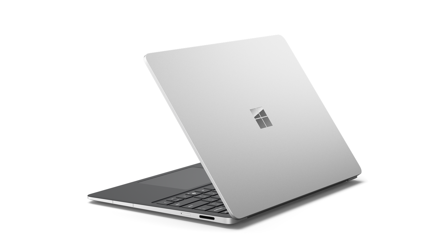 Laptop plateado abierto en ángulo. Logotipo de Microsoft en la tapa. Teclado visible. Fondo gris.