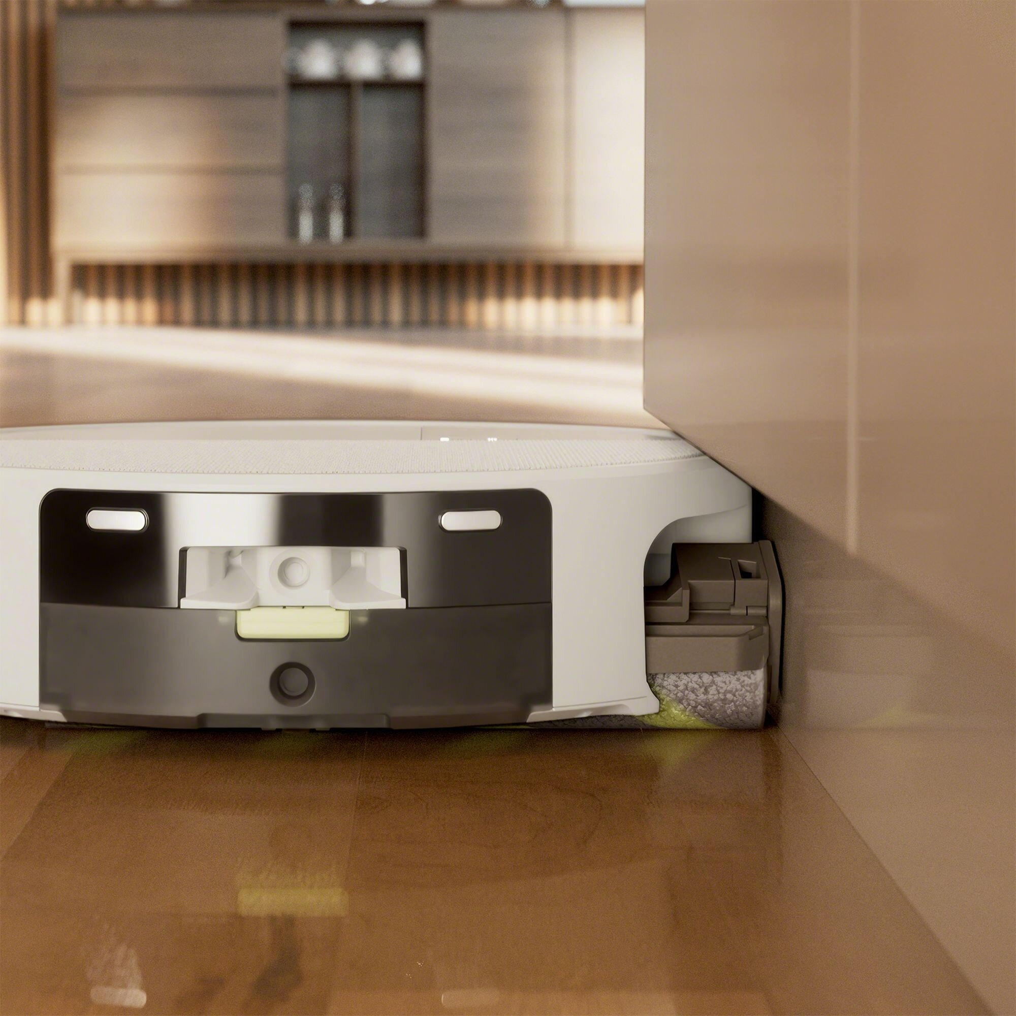 Robot odkurzający jest blisko ściany. Podłoga jest brązowa. Odkurzacz jest biały i czarny.