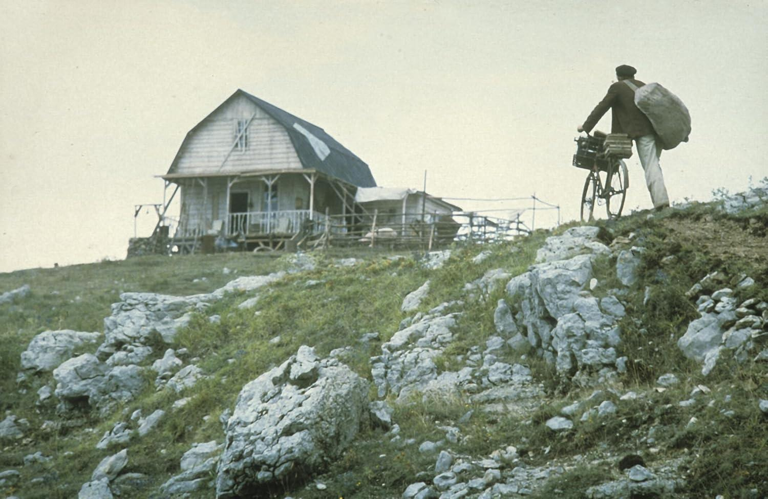 Ein Mann mit einem Sack auf einem Fahrrad erklimmt einen Hügel. Auf dem Hügel steht ein Haus. Felsen und Gras befinden sich im Vordergrund.