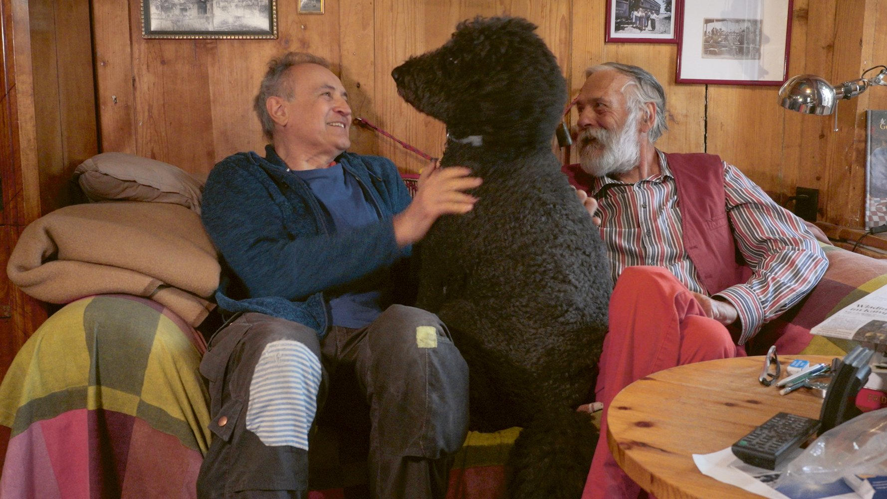 Zwei Männer und ein großer schwarzer Hund sitzen auf einem Sofa. Ein Mann streichelt den Hund. Holzwände und gerahmte Bilder sind im Hintergrund.