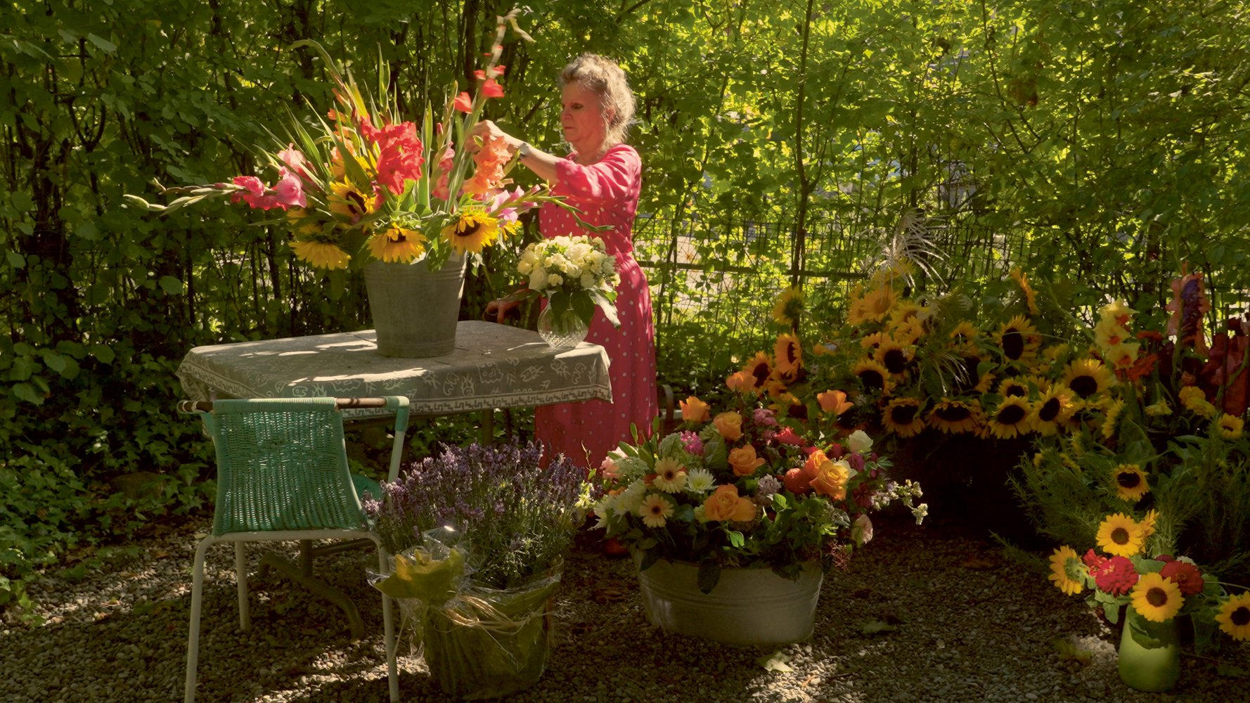 Eine Frau in einem rosa Kleid arrangiert Blumen in einem Garten voller Sonnenblumen, Rosen und anderer Blüten.