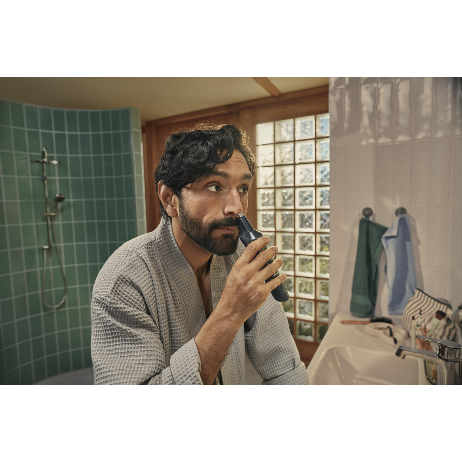 A man shaves his beard in a bathroom. He wears a robe. A sink, mirror, and shower are visible in the background.