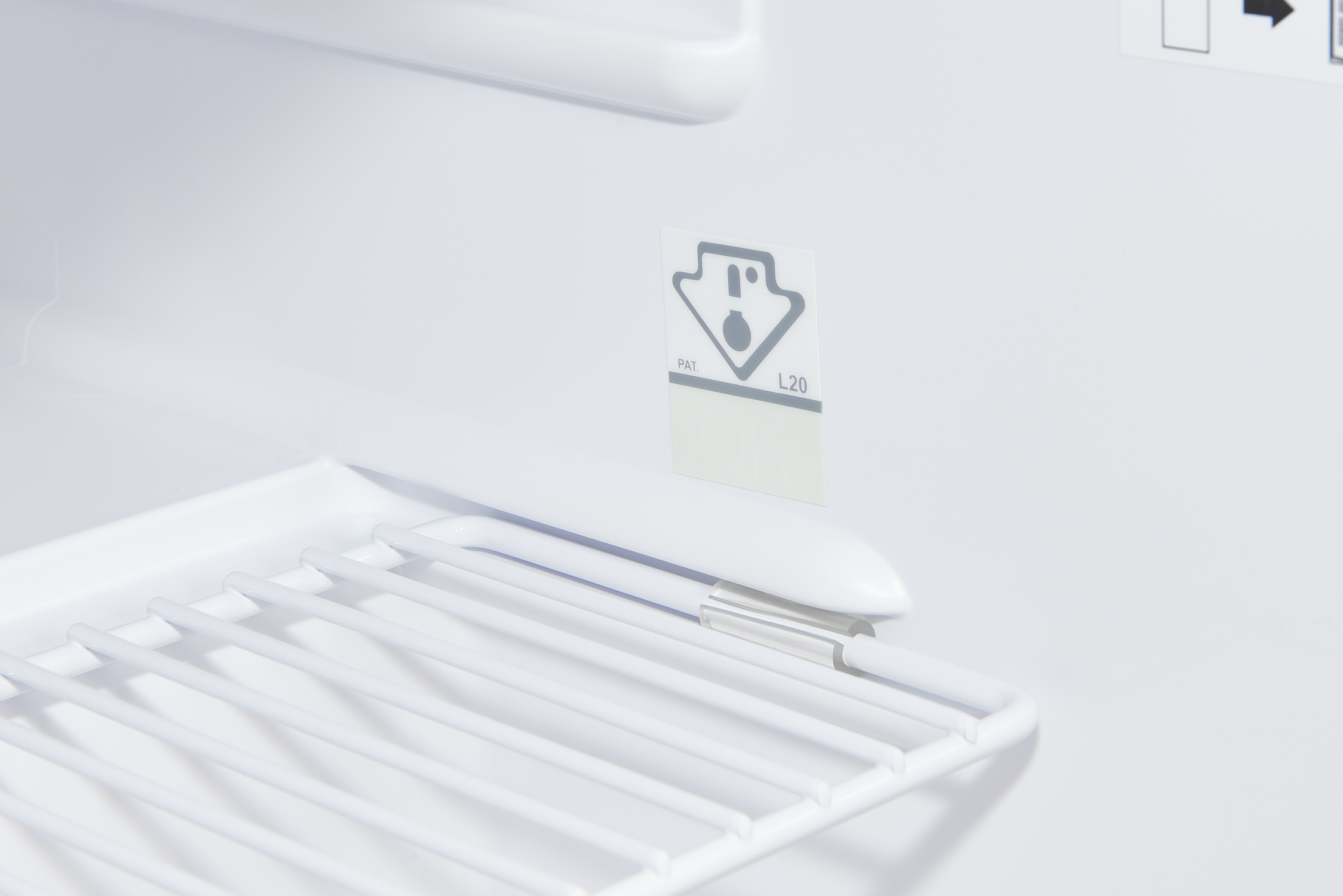 À l'intérieur d'un réfrigérateur blanc. Une étagère en fil blanc en bas à gauche. Un autocollant avec un point d'exclamation est au centre.