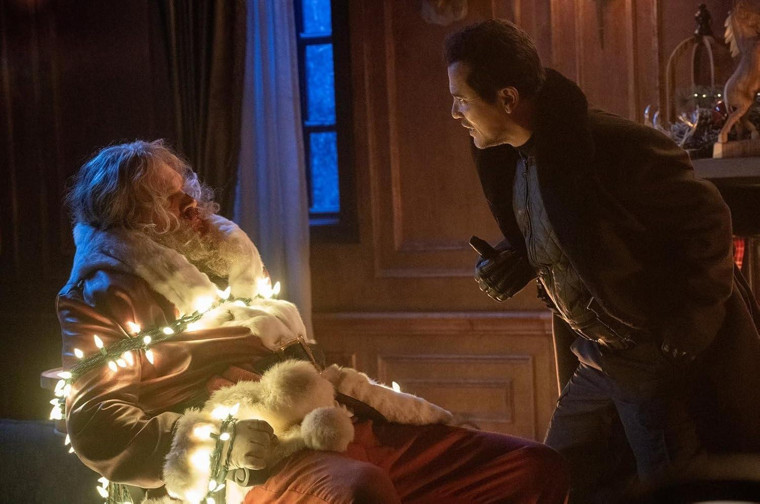 Der Weihnachtsmann, in Lichterketten gehüllt, sitzt einem Mann in Schwarz gegenüber, mit ernstem Blick, in einem schwach beleuchteten Raum.