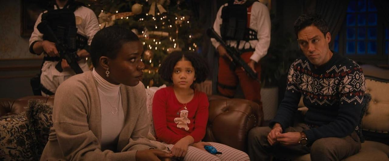 Familie, Kind und Mann sitzen auf einer Couch in der Nähe eines Weihnachtsbaums. Bewaffnete Männer stehen im Hintergrund. Die Szene wirkt angespannt.