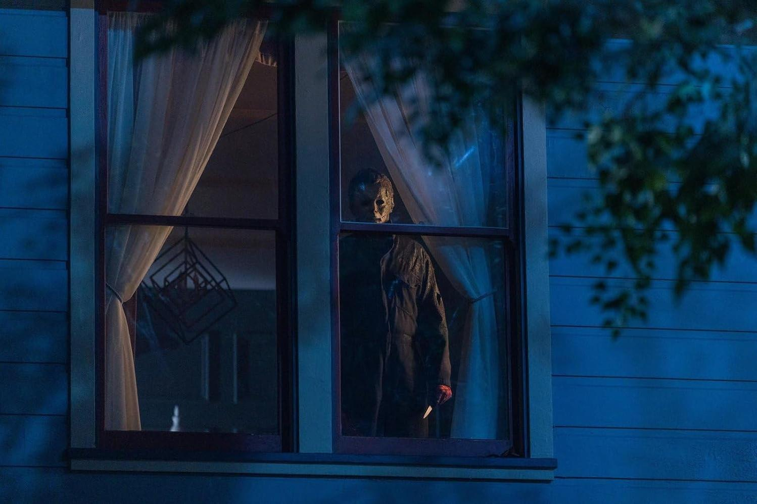 Eine maskierte Person steht hinter einem Fenster und hält ein Messer. Das Fenster wird von einem blauen Haus mit Vorhängen und einer Hängedekoration umrahmt.