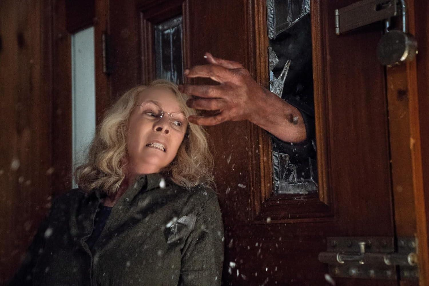 Eine Frau reagiert auf eine Hand, die durch zerbrochenes Glas in einer Tür greift. Die Hand ist dunkel, die Frau trägt eine Brille und ein grünes Hemd.