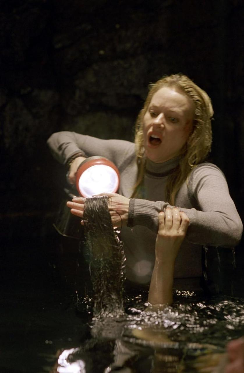 Frau im Wasser hält eine leuchtende Taschenlampe, Haar in der Hand, sieht verängstigt aus. Ein weiterer Arm taucht aus dem Wasser auf. Dunkler, höhlenartiger Hintergrund.