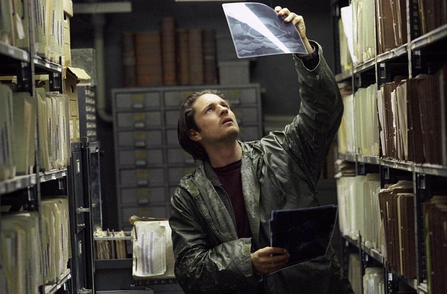 Ein Mann in einer Bibliothek betrachtet ein Dokument, umgeben von Regalen voller Akten. Er trägt eine Jacke und blickt nach oben, beleuchtet von oben.