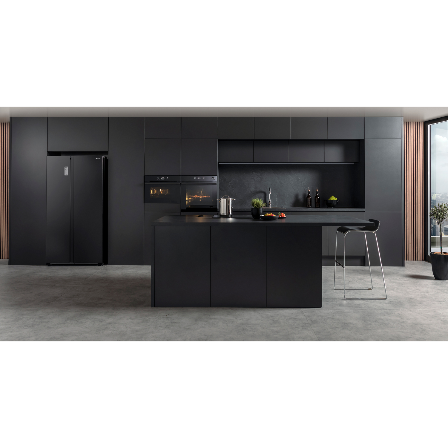 Modern black kitchen with refrigerator, ovens, island, and bar stool. Gray floors and a large window with a city view.