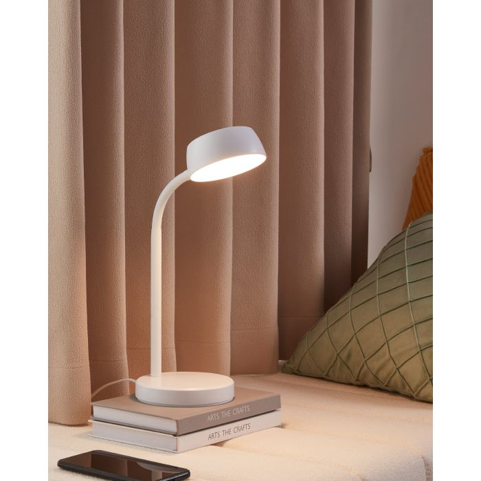 Eine weiße Schreibtischlampe steht auf Büchern, davor ein Telefon. Im Hintergrund Vorhänge und rechts ein Kissen.