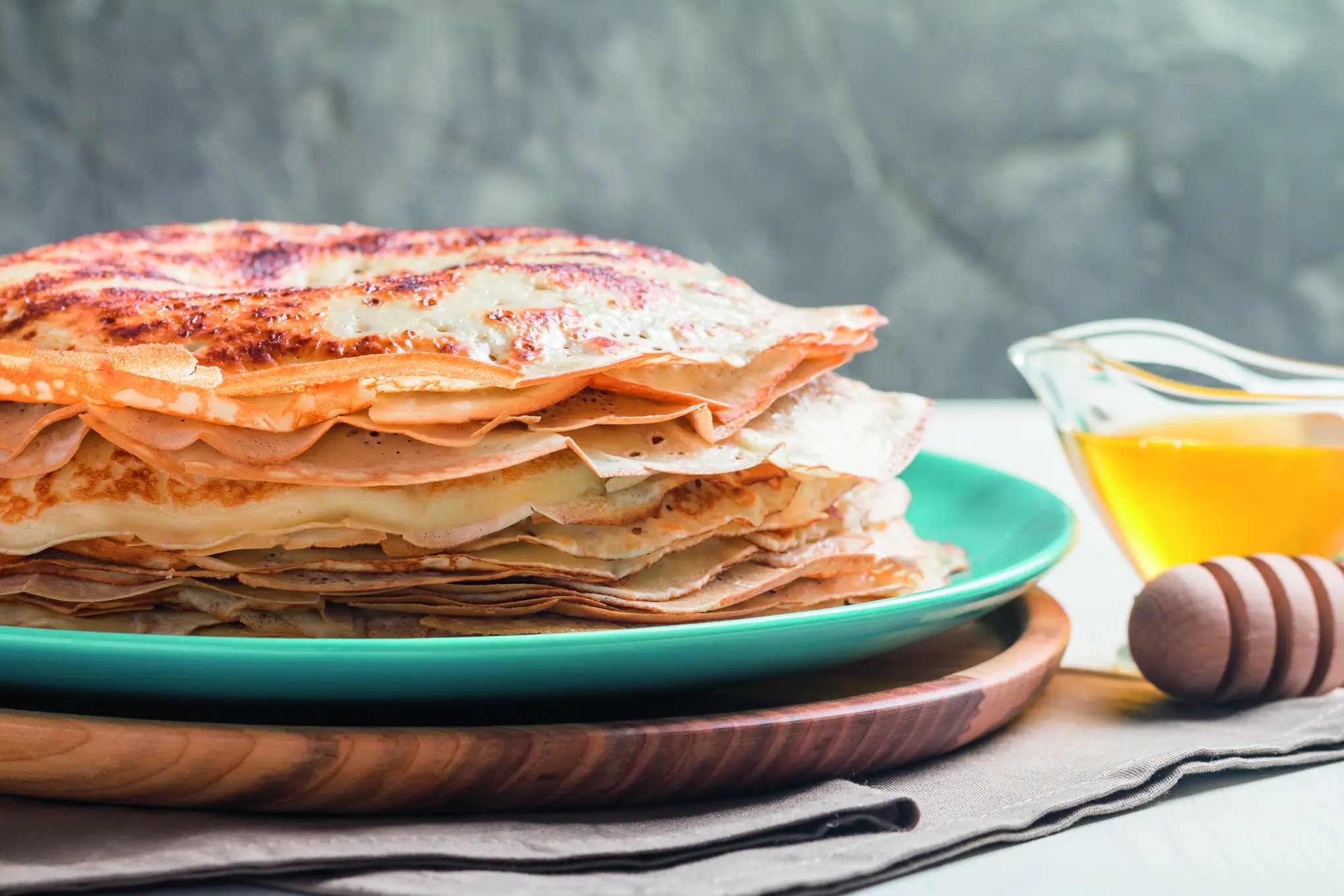 Ein Stapel goldener Pfannkuchen auf einem türkisfarbenen Teller, einem Holztablett, mit Honig und einem Honiglöffel, alles auf einer grauen Tischdecke.