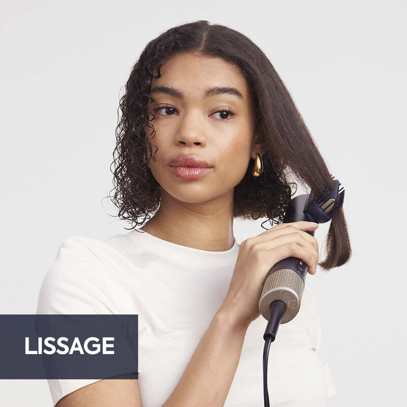 Eine Frau mit lockigem Haar benutzt ein Haarstyling-Tool. Das Werkzeug glättet einen Teil ihres Haares. Sie trägt ein weißes Hemd und goldene Ohrringe.