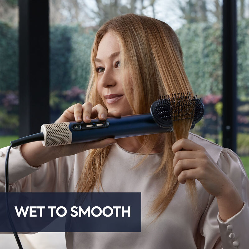 Blonde Frau stylt ihr Haar mit einem blau-schwarzen Bürsten-Haartrockner. Der Text lautet 'WET TO SMOOTH'.