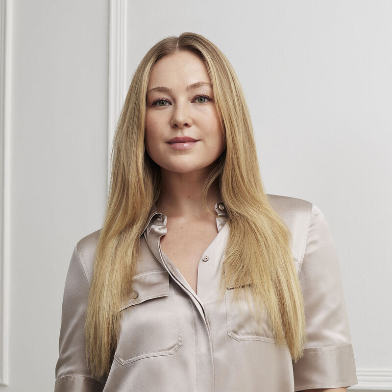 Eine hellhäutige Frau mit langen blonden Haaren und einem Seidenhemd lächelt. Der Hintergrund ist eine weiße Wand.