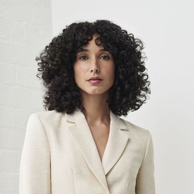 Eine Frau mit lockigem, schwarzem Haar trägt einen cremefarbenen Blazer. Sie schaut direkt in die Kamera. Der Hintergrund ist eine weiße Ziegelmauer.
