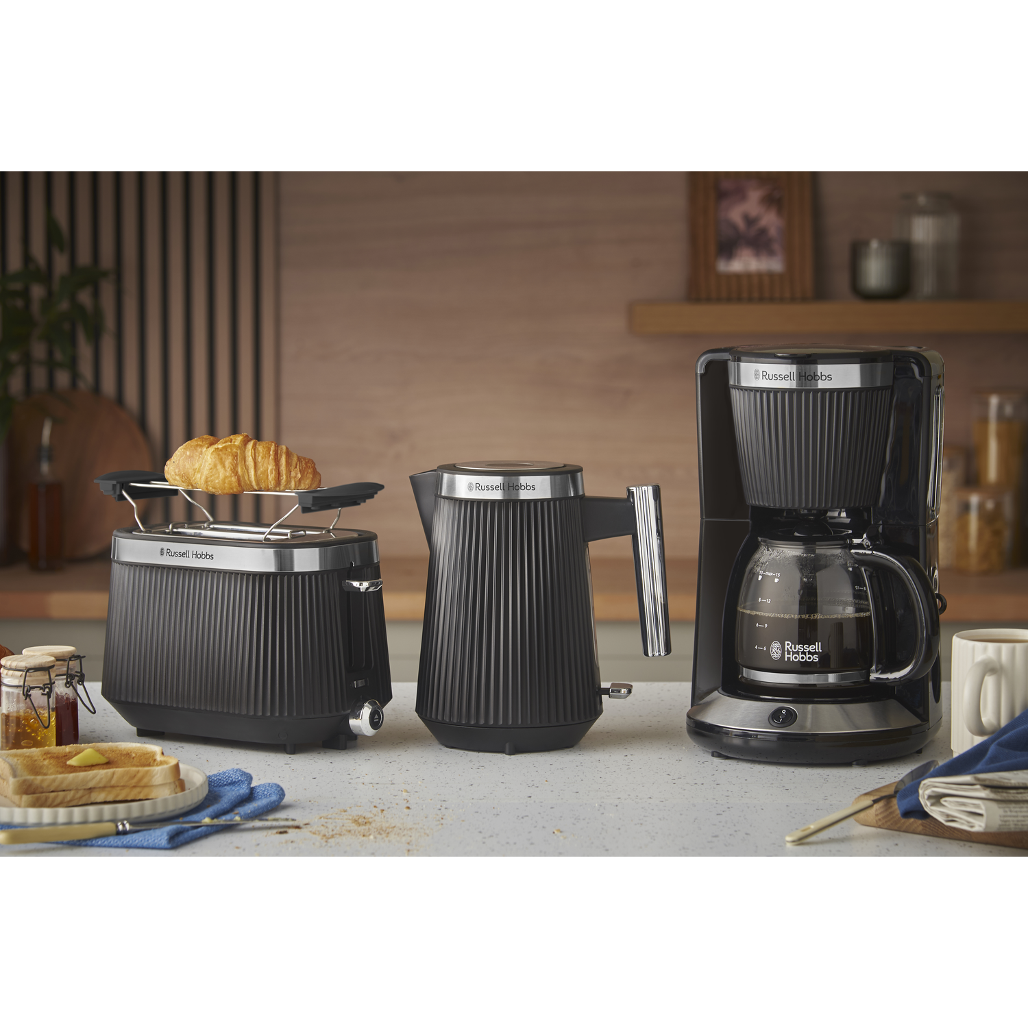 A kitchen scene with black Russell Hobbs appliances: a toaster with a croissant, a kettle, and a coffee maker. Toast and honey are on a plate.