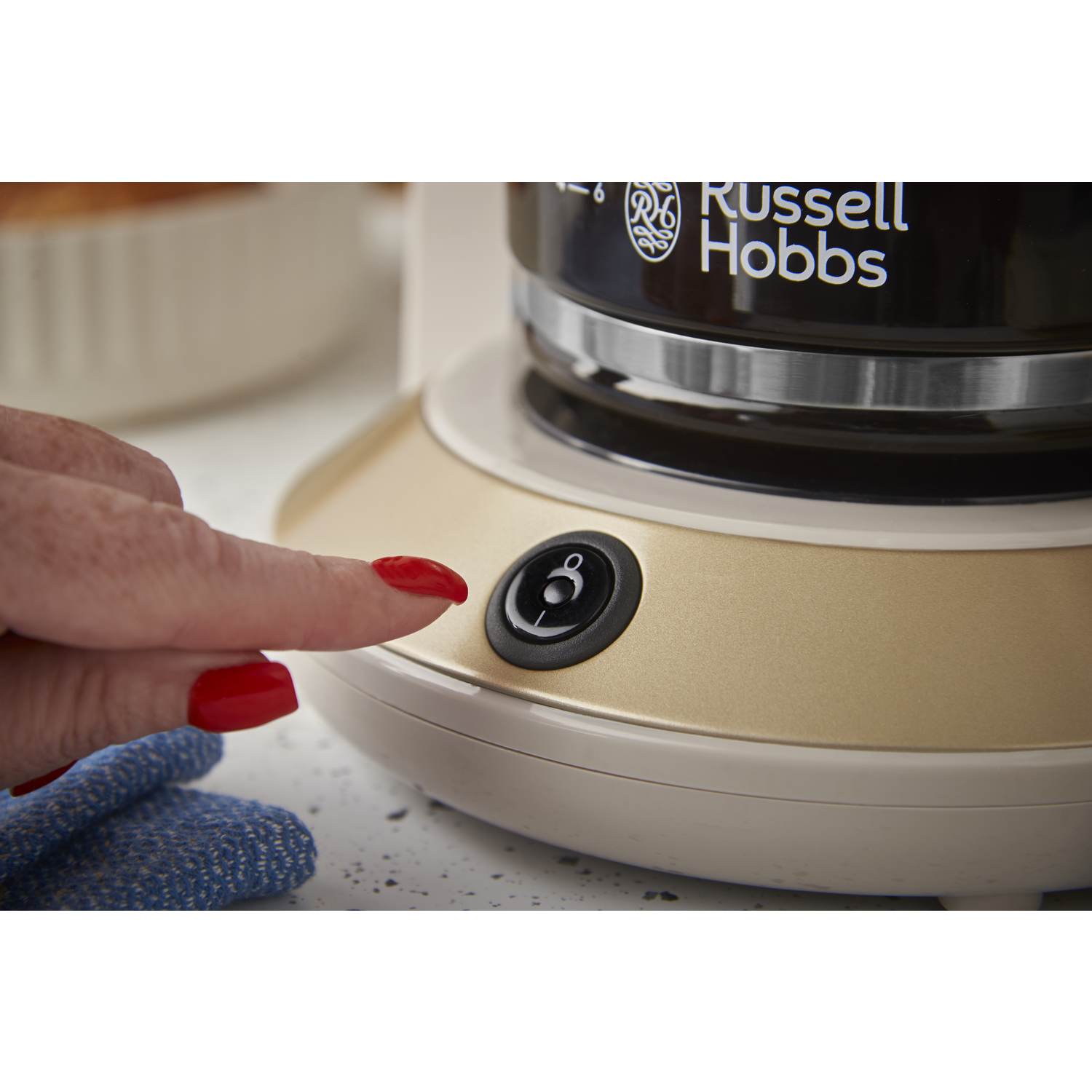 A hand with red nails presses a black power button on a gold and cream coffee maker. A blue cloth is next to it.